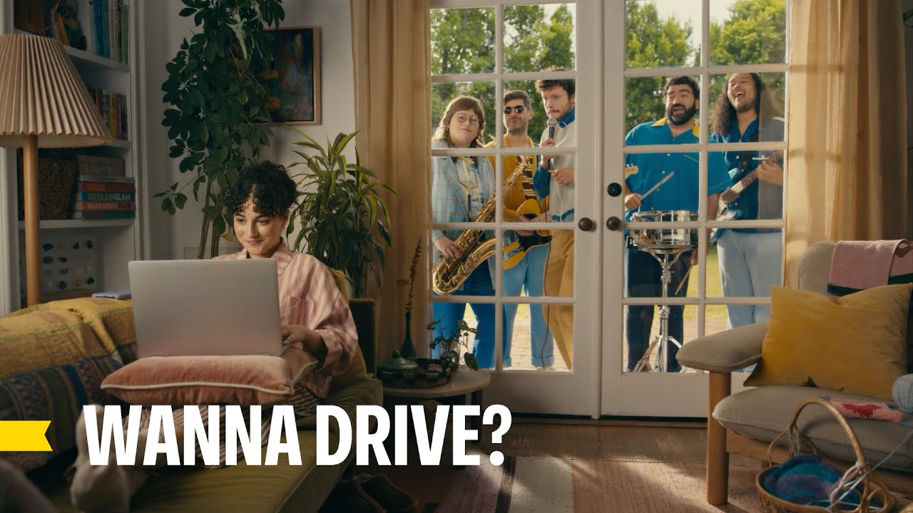 Still from CarMax's Wanna Buy Your Way campaign showing a shopper using a laptop while a band waits outside